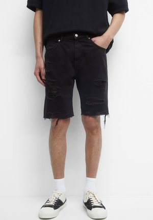 Personne portant un short en jean noir déchiré, des chaussettes blanches, des baskets noires et blanches, debout avec une main dans la poche, sur fond blanc.