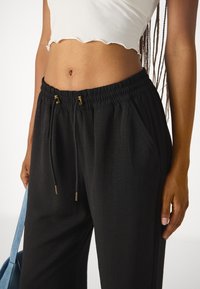 Black drawstring pants with an elastic waistband, featuring side pockets and a textured fabric. Gold hardware accents on drawstrings.