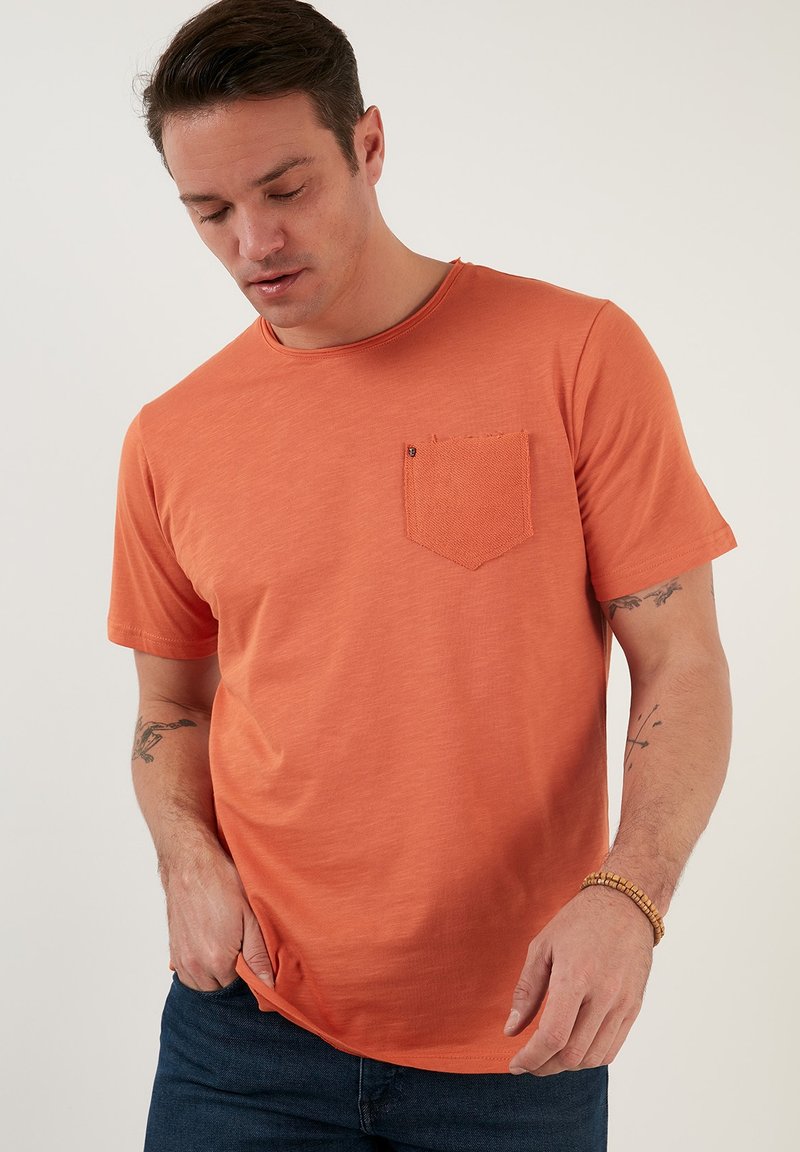 Krátkoročni oranžni T-shirt iz mehkega blaga, z teksturirano prsno žepom in okroglo ovratnikom, oblečen z temno modrimi kavbojkami.