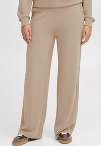 Beige, vide bukser lavet af blødt stof med elastisk talje. Har en glat tekstur og et afslappet design. Sammen med brune sneakers.