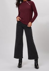 Pull côtelé bordeaux avec trois boutons décoratifs sur l'épaule, associé à un jean noir à jambes larges et des bottines.