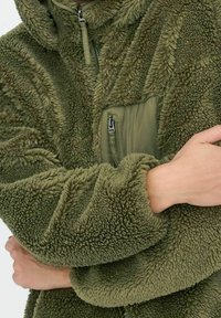 Chlupatá zelená fleecová bunda se zipovou kapsou vpředu, texturovanou tkaninou, oversized kapucí a širokými rukávy. Měkký, teplý a neformální design.