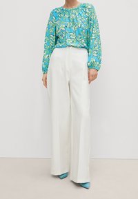 Blouse à imprimé floral en bleu, vert et blanc ; manches longues avec fronces. Pantalons blancs à jambes larges avec une texture lisse. Chaussures à talons hauts bleues.