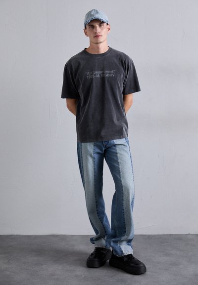 Junger Mann, der ein verblasstes schwarzes T-Shirt mit der Aufschrift "Art Department", zweifarbige blaue Jeans, schwarze Sneaker und eine hellblaue Jeansmütze trägt, steht vor einer grauen Wand.
