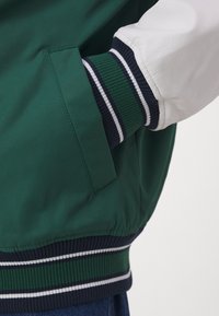Casaco verde com mangas brancas, apresentando punhos canelados azul marinho com riscas brancas e um bolso lateral. Textura suave, design desportivo.