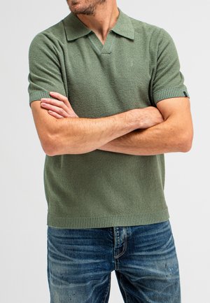 Short-sleeved polo shirt in a soft, textured olive green fabric with a ribbed collar and cuff details, paired with distressed blue jeans.