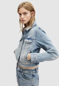 Giacca in denim azzurro chiaro tagliata sopra la vita con chiusura a bottoni, tasche sul petto e dettagli strappati sui bordi. Abbinata a jeans in denim coordinati.