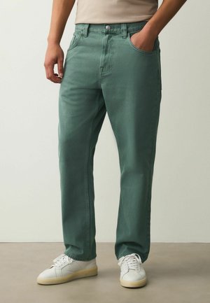 Mann trägt grüne gerade geschnittene Hose und weiße Sneakers, steht mit einer Hand in der Tasche vor einfarbigem Hintergrund.