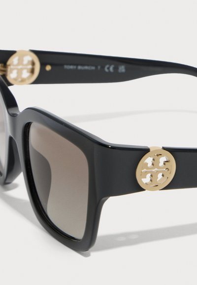 Γυαλιά ηλίου με μαύρους τετράγωνους σκελετούς, φακούς με απόχρωση βαθμιδωτής μετάβασης και χρυσή στρογγυλή λεπτομέρεια λογότυπου στο μπράτσο του βραχίονα, τοποθετημένα σε λευκό φόντο.