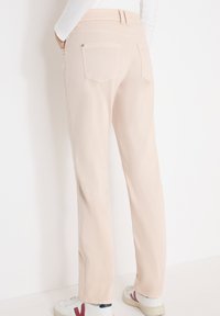 Personne portant un pantalon beige à jambes droites et des baskets blanches avec des accents rouges, debout devant un fond blanc uni.