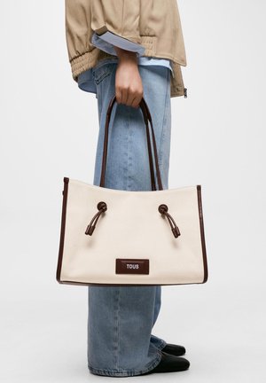 Persona que lleva una chaqueta beige y vaqueros azules sosteniendo un bolso de lona blanco con asas marrón oscuro y el logo de TOUS.