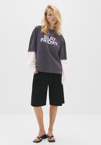 Lilla oversized t-shirt med hvid og pink "SLAY FRIDAY" tekst, lagd ovenpå en hvid langærmet skjorte, parret med sorte shorts og sorte sandaler.