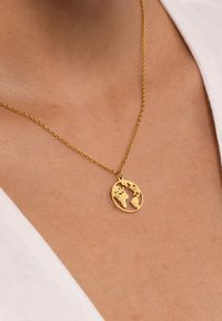 Goldene Halskette mit einem kreisförmigen Anhänger im Design einer ausgeschnittenen Weltkugel, aufgehängt an einer filigranen Kette, die über dem Schlüsselbein ruht.