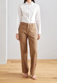 Chemise blanche boutonnée à manches longues, associée à un pantalon beige tailleur. Dispose d'un col classique, d'un devant plat et d'une coupe décontractée.