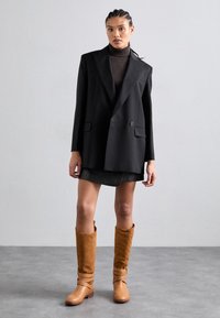 Blazer noir à double boutonnage porté sur un col roulé marron foncé, associé à une jupe gris foncé et des bottes hauteur genou de couleur marron clair avec des sangles.