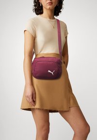 Puma crossbody taske i mørk bordeaux, lavet af blødt nylon, med en frontlomme med lynlås og ikonisk hvidt logo. Justerbar rem inkluderet.