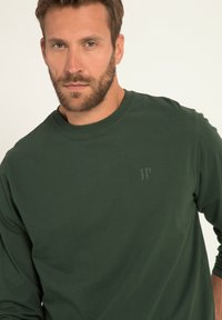 Dunkelgrünes Langarmshirt mit Rundhalsausschnitt. Verfügt über ein dezentes Logo auf der Brust. Glatte Textur und figurbetontes Design.