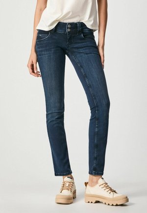 Mujer con vaqueros pitillo azul oscuro con cierre de doble botón y zapatillas beige con suela gruesa, de pie frente a un fondo liso.
