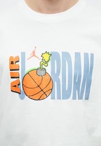 Λευκό μπλουζάκι με σχέδιο γραφικών που περιλαμβάνει το κείμενο "AIR JORDAN", μια πορτοκαλί μπάλα του μπάσκετ με φυτίλι και πράσινες λεπτομέρειες, και μπλε γράμματα.