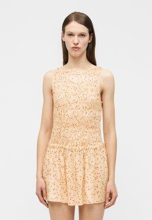 Fiatal nő egyenes barna hajjal, ujjatlan bézs ruhában narancssárga pöttyökkel, egyszínű világos háttér előtt állva.