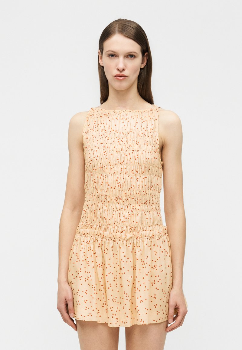 Νεαρή γυναίκα με ίσια καστανά μαλλιά που φοράει αμάνικο μπεζ φόρεμα με πορτοκαλί πουά, όρθια μπροστά σε απλό ανοιχτό φόντο.