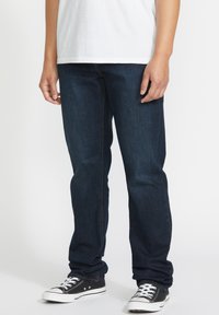 Jeans de mezclilla azul oscuro con corte recto, con costuras naranjas y una textura suave, combinados con zapatillas negras.