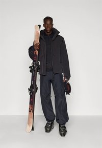 Smučarska oprema vključuje črno jakno, črne hlače, smučarske čevlje in čelado. Smučar drži lesene smuči z temnimi vezmi.