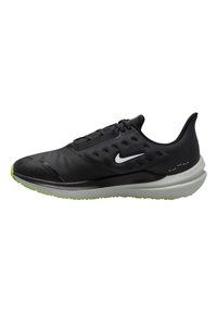 Zapatilla deportiva negra con parte superior suave, sistema de cordones y suela negra texturizada. Presenta una suela verde y el logo de Nike reflectante.