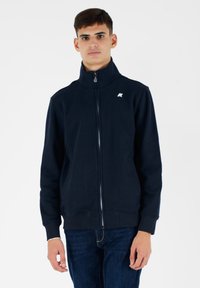 Giacca zip-up navy con colletto alto, realizzata in morbido tessuto. Presenta un logo bianco in alto a sinistra e due tasche laterali. Texture liscia.