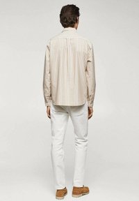 Camisa de rayas beige de manga larga con dobladillo redondeado, combinada con jeans blancos y zapatos marrones. Textura suave y corte clásico.