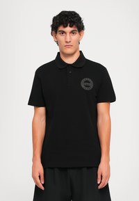 Bărbat îmbrăcat cu un polo negru cu mânecă scurtă, având un mic logo rotund "Versace Jeans Couture" pe partea stângă a pieptului, stând în fața unui fundal simplu.