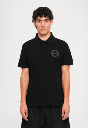 Mann iført svart kortermet polo-skjorte med en liten rund "Versace Jeans Couture"-logo på venstre bryst, som står foran en ensfarget bakgrunn.