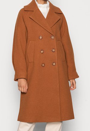 Long manteau en laine marron à double boutonnage avec larges revers et poches avant, porté sur un pull blanc et un pantalon beige.