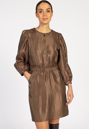 Vrouw in een glanzende bruine jurk met een riem, met opgestoken lange mouwen en een rits aan de voorkant, staand met één hand in de zak tegen een witte achtergrond.