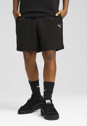 Person iført sorte Puma-shorts, sorte Puma-strømper og sorte Puma-sneakers, stående med hænderne i lommerne på en grå baggrund.