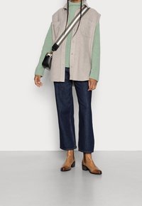 Beige mouwloze vest over een groene geribbelde trui, donkerblauwe wijde jeans en bruine enkellaarzen. De outfit heeft minimalistische details.