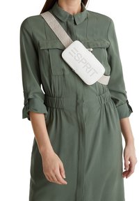Bolso cruzado de cuero blanco con el logo "ESPRIT" en relieve, sujeto a una correa estampada en beige, llevado sobre un vestido de camisa verde.