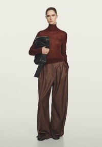 Brązowy sweter typu golf, nałożony na wysokoprocentowe, szerokie spodnie w kratę; modelka trzyma czarną skórzaną torbę. Gładka tekstura, nowoczesny krój.