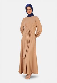 Beige long dress with a knotted waist and long sleeves, made of lightweight fabric. Paired with a blue hijab and strappy sandals.