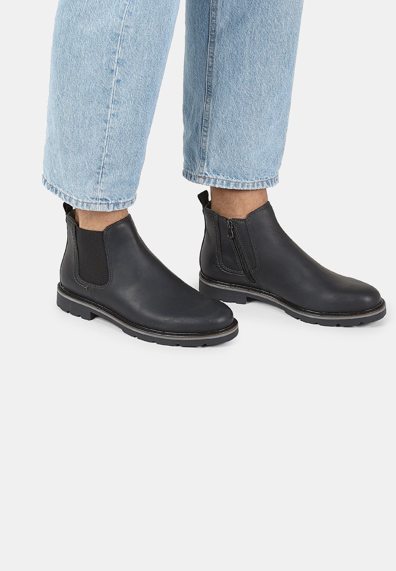 Stivali neri in pelle alla caviglia con pannelli laterali elastici e chiusura con zip, superficie testurizzata, punta arrotondata e dettagli sul suolo in contrasto.