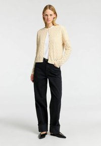 Cardigan lavorato a maglia color crema con motivo a rombi, apertura frontale e bordi a costine, indossato sopra una camicia bianca e abbinato a jeans scuri e mules neri.