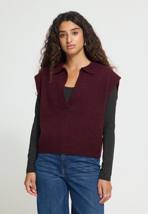 Femme aux longs cheveux bouclés portant un gilet tricoté bordeaux sans manches sur un haut noir à manches longues et un jean bleu, face à l'avant.