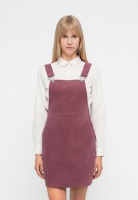 Vestido de tirantes de pana en color malva apagado, con tirantes ajustables, bolsillos frontales y textura acanalada, combinado con una camisa blanca con cuello.