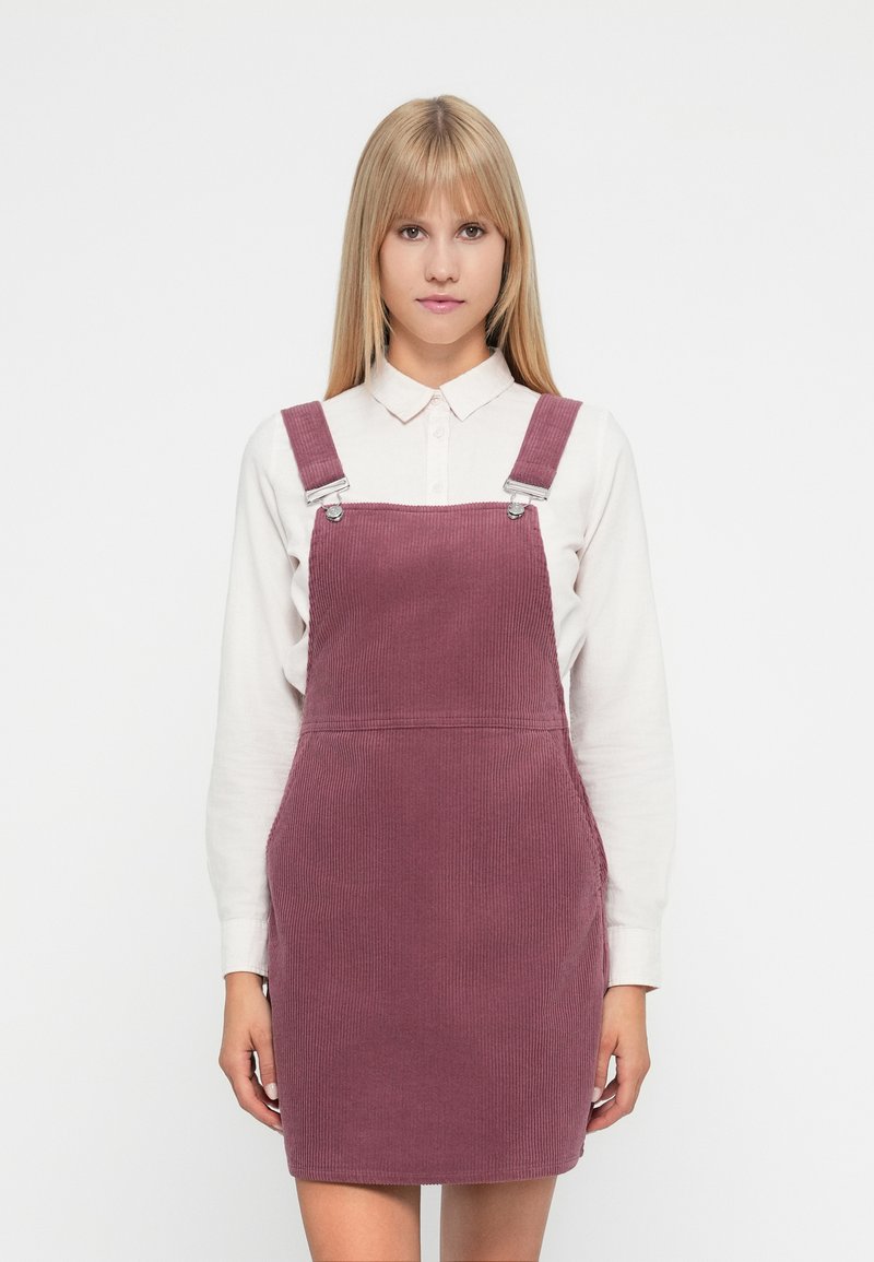 Vestido de tirantes de pana en color malva apagado, con tirantes ajustables, bolsillos frontales y textura acanalada, combinado con una camisa blanca con cuello.