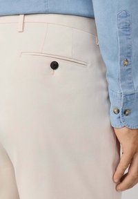 Kamera u bližem planu prikazuje bež hlače s crnom dugmadi na stražnjem džepu, u kombinaciji s plavom košuljom dugih rukava čiji je manžet vidljiv blizu ruke.