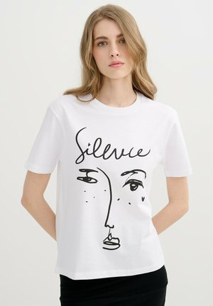 Joven mujer con largo cabello rubio que lleva una camiseta blanca con una cara abstracta negra y la palabra "Silvice" escrita encima.