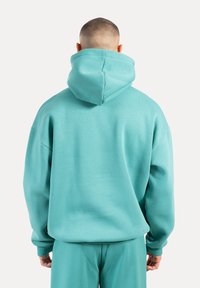 Hoodie turquoise surdimensionné avec capuche, fabriqué à partir d'un matériau doux ; présente des épaules tombantes et des poignets côtelés. La vue de dos met en avant la coupe décontractée.