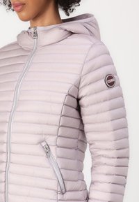 Giacca a sacco di colore viola chiaro con cappuccio, design trapuntato, zip frontale e tasca laterale. Presenta un logo circolare del marchio sulla manica.