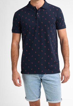Mand iført en navyblå polo med små røde mønstre og lyseblå denimshorts med rullede kanter.