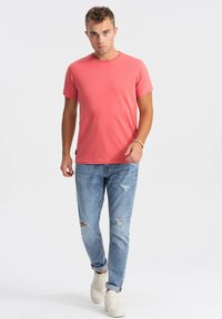 Koralfarvet t-shirt med korte ærmer lavet af bomuld, parret med slidte lysblå jeans og hvide sneakers. Model står med en neutral udtryk.